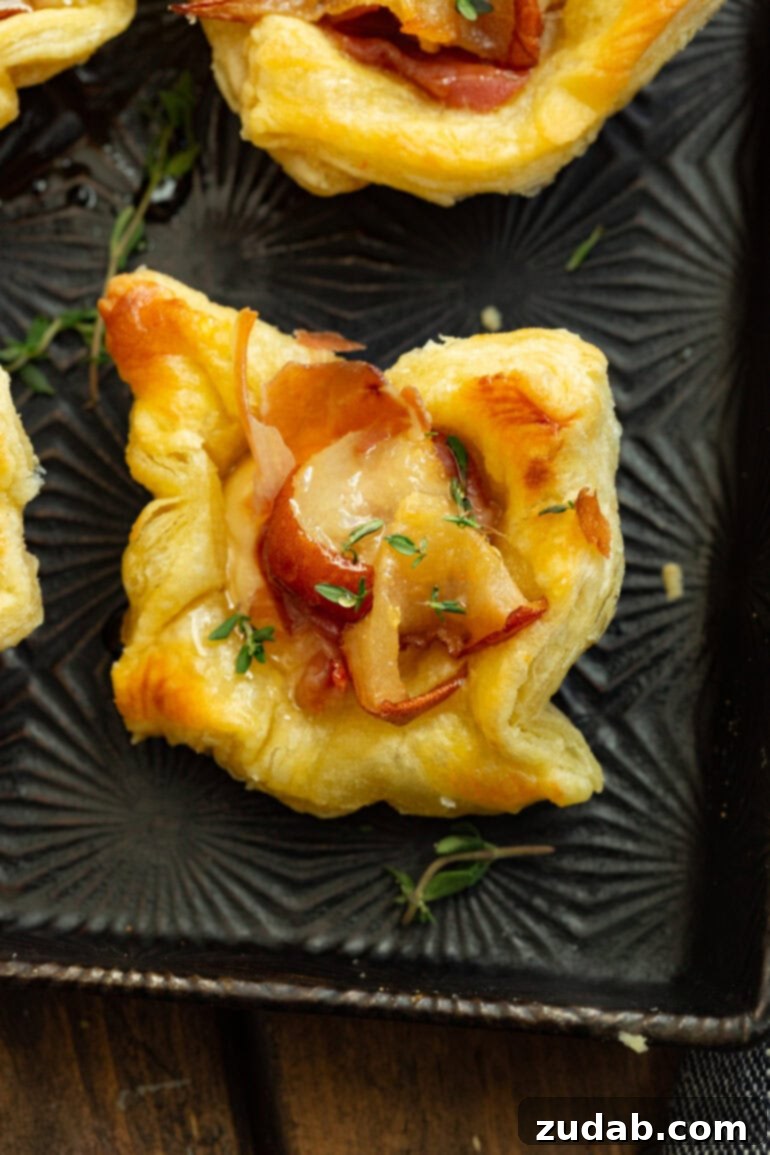 An overhead shot showcasing a single, perfectly golden-baked puff pastry bite, overflowing with roasted pear, melted brie, a curl of prosciutto, and garnished with a sprig of fresh thyme.