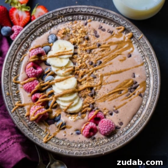 Weekly Menu Plan for January 8 to 14 6 A Kid-Friendly Chocolate Banana Smoothie Bowl, beautifully arranged on a black board. The bowl itself is vintage silver, nestled next to a purple napkin and two vintage silver spoons. The smoothie is generously topped with fresh raspberries, sliced bananas, blueberries, crunchy granola, and chocolate chips, all elegantly drizzled with creamy peanut butter. A glass of milk sits invitingly behind it.