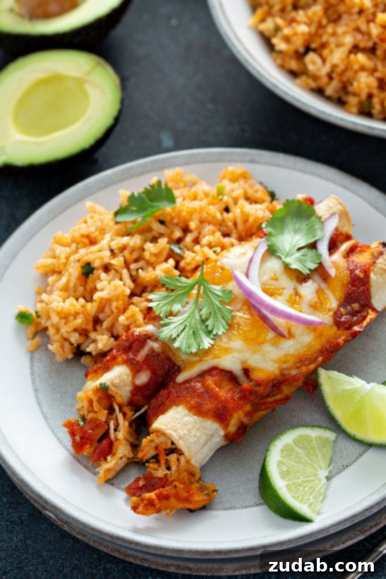 A dinner plate with two red chicken enchiladas, two lime wedges, and a serving of Spanish rice. The enchiladas have red sauce, melted cheese, slices of red onion, and cilantro leaves on top. There is a avocado that is sliced in half and a bowl of Spanish rice in the background. A festive meal for Friday.