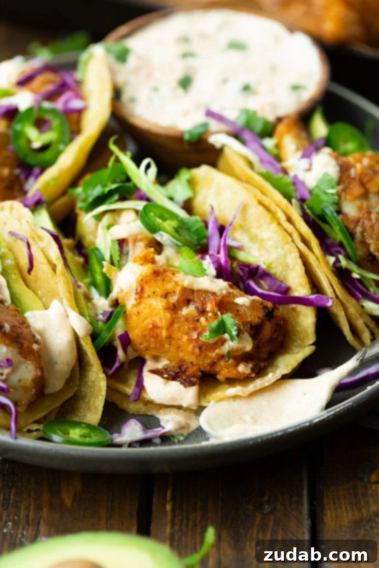 Effortless Homemade Fish Tacos 4 a photo of several fish tacos sitting side by side on a plate topped with shredded cabbage, sliced avocados and jalapenos and a creamy sauce