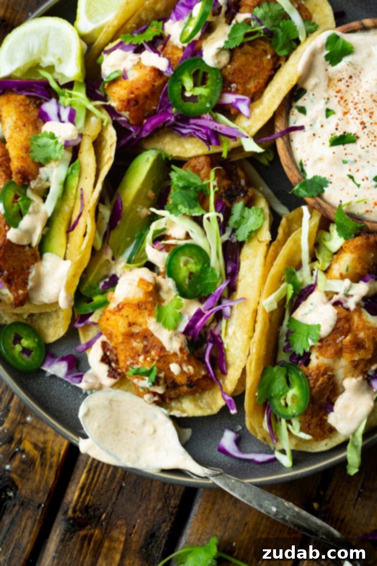 Effortless Homemade Fish Tacos 5 a photo of several homemade fish tacos sitting next to each other on a plate topped with sliced jalapenos, avocados, shredded green and purple cabbage and white fish taco sauce