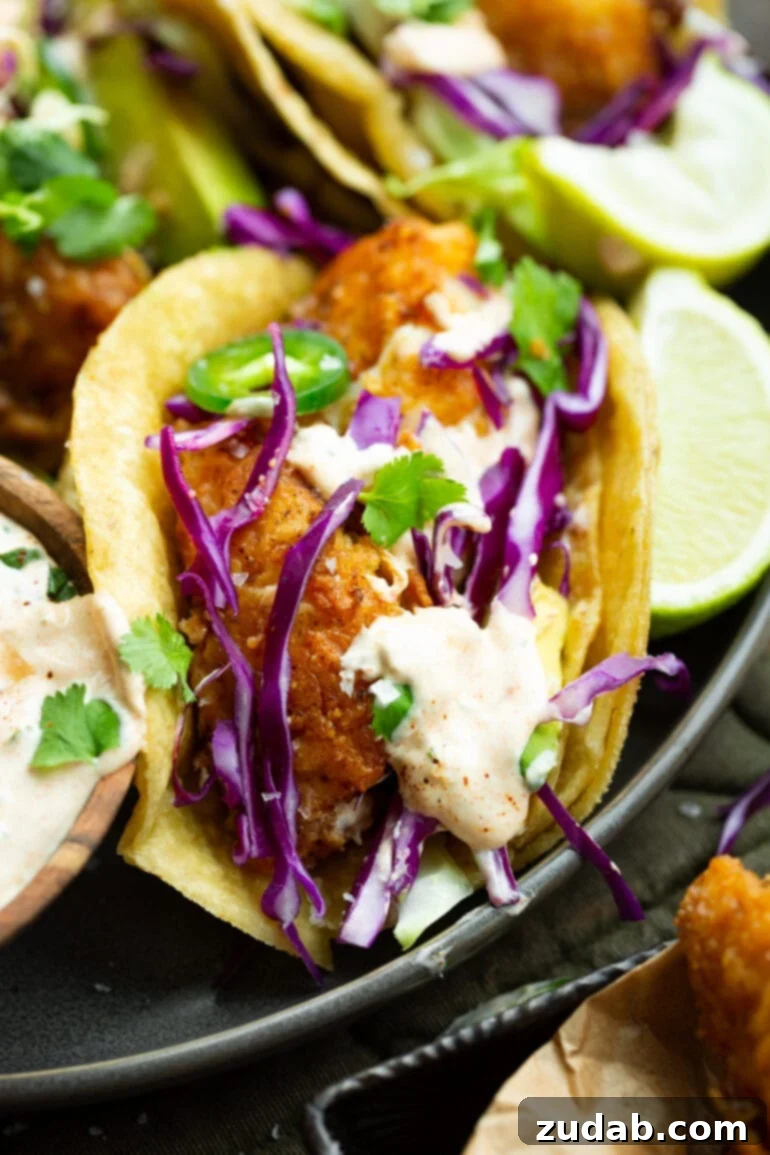 Effortless Homemade Fish Tacos 8 a photo of a golden pan fried piece of fish in a corn tortilla topped with shredded cabbage, sliced jalapenos and a creamy sauce.