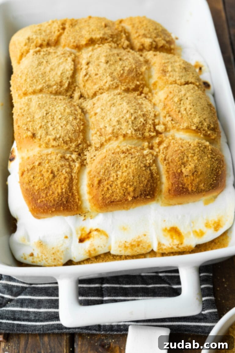 Gooey Smores Sliders 3 A casserole dish brimming with a dozen golden-brown s'mores sliders, showcasing deliciously melted marshmallow and chocolate spilling out, all crowned with a generous sprinkle of graham cracker crumbs.