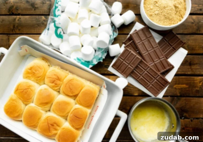 Gooey Smores Sliders 4 A flat lay photograph showcasing all the simple ingredients required for s'mores sliders: a package of Hawaiian sweet rolls, a bowl of melted butter, a bag of large marshmallows, several Hershey's chocolate bars, and a bowl of graham cracker crumbs.