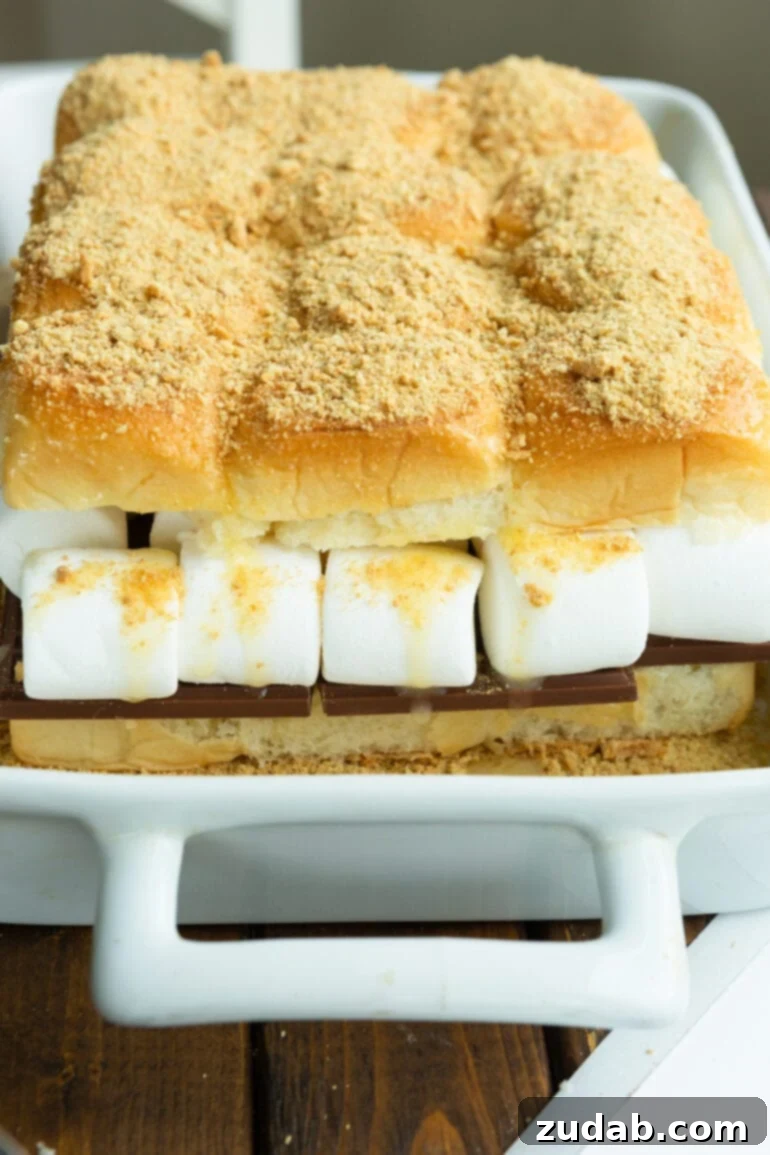 Gooey Smores Sliders 6 A baking dish filled with s'mores slider buns, generously layered with chocolate and large marshmallows, then topped with graham cracker crumbs, poised and ready to be baked into a golden, gooey dessert.