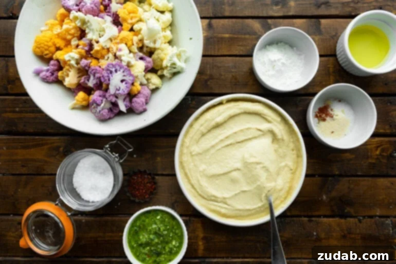 Crispy Roasted Cauliflower with Creamy Hummus 3 a photo of all the ingredients for roasted cauliflower including a bowl of hummus, a small bowl of pesto sauce and a plate of orange and purple cauliflower florets