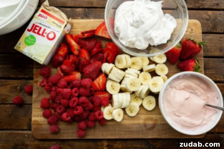 Fluffy Cheesecake Fruit Salad 4 a photo of all the ingredients sitting on a wooden cutting board including sliced bananas, strawberries and whole raspberries with bowls of strawberry yogurt, a box of cheesecake jello and whipped cream next to it