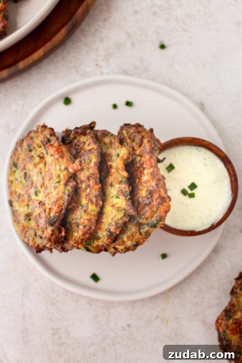 a photo of golden crispy zucchini fritters standing up on a plate leaning up against a small cup of dipping sauce
