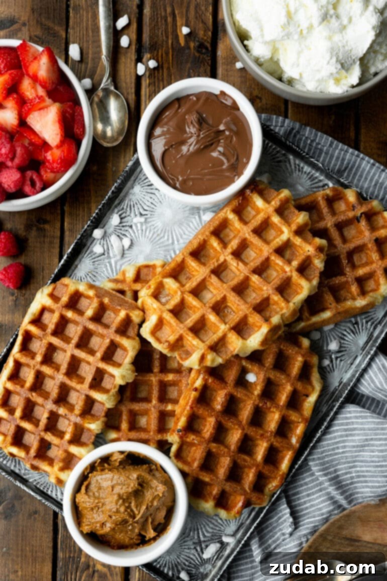 Mastering Waffle Love's Signature Liege Waffles 7 A photo of several golden cooked Liege waffles piled on a baking sheet with pearl sugar scattered around them and small bowls of Nutella and Biscoff sitting next to them, ready to be topped.