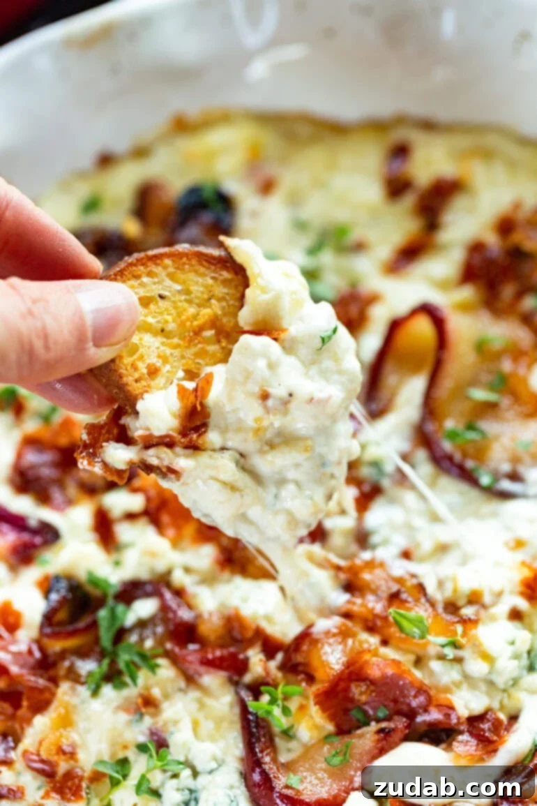 Sweet Pear, Savory Prosciutto & Tangy Blue Cheese Dip 3 A close-up photo of a hand dipping a crostini into a dish full of gooey roasted pear and blue cheese dip, showcasing its creamy texture.