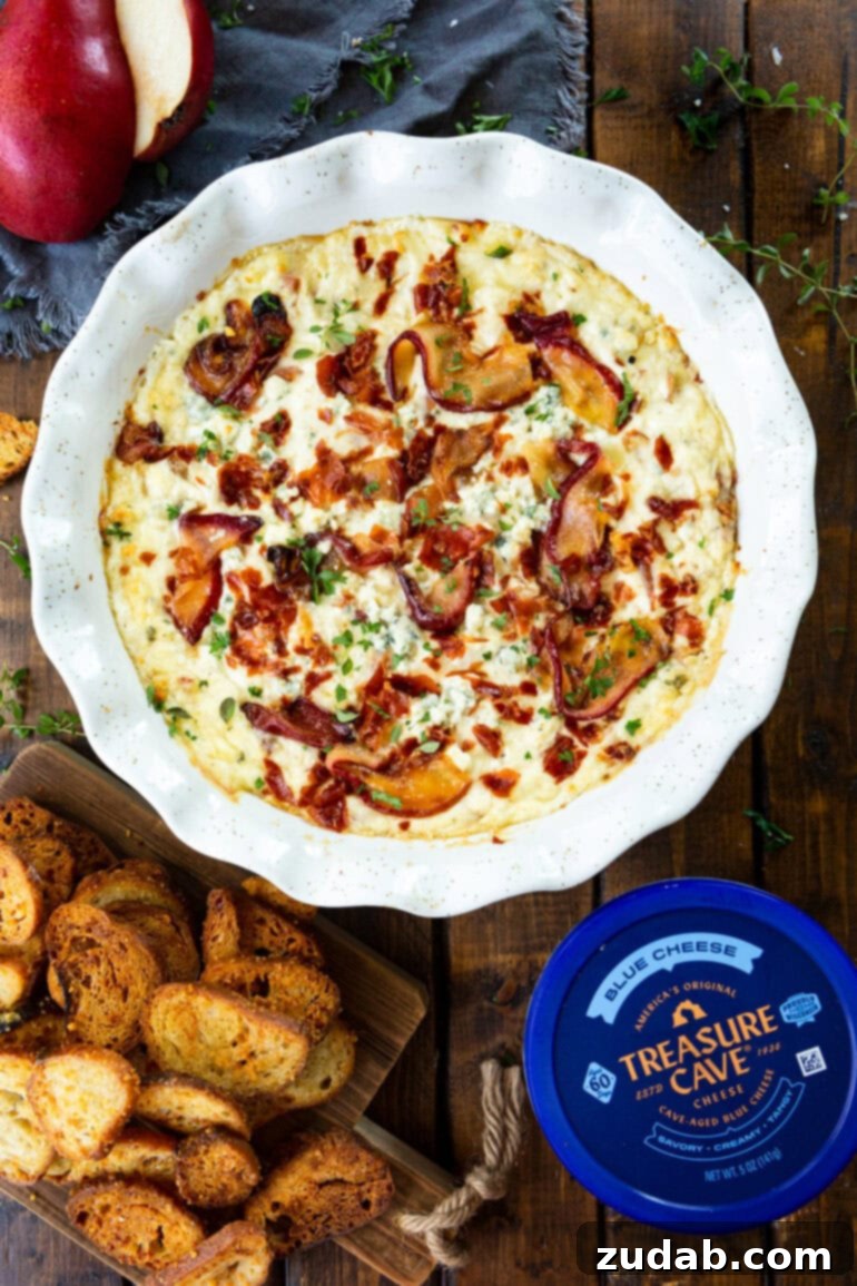 Sweet Pear, Savory Prosciutto & Tangy Blue Cheese Dip 5 A dish of baked blue cheese dip topped with roasted pears and crispy prosciutto, with a container of Treasure Cave blue cheese next to it on a rustic wooden surface.