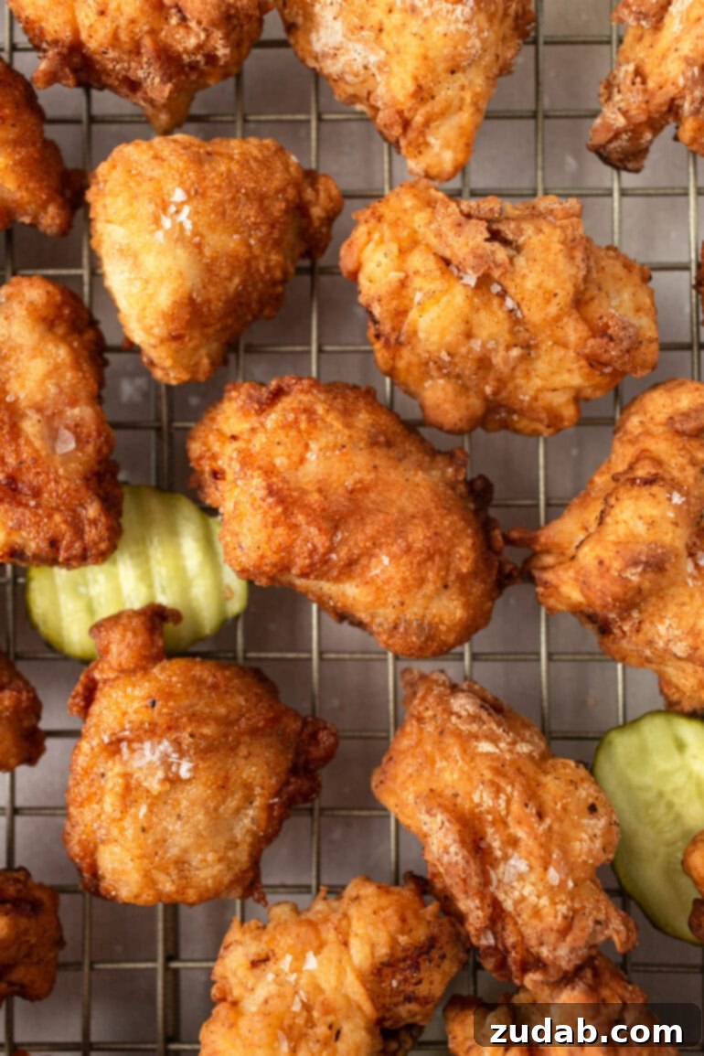 A close-up shot of several golden, deep-fried chicken nuggets resting on a wire cooling rack, with a few dill pickle chips scattered around, hinting at the secret ingredient.