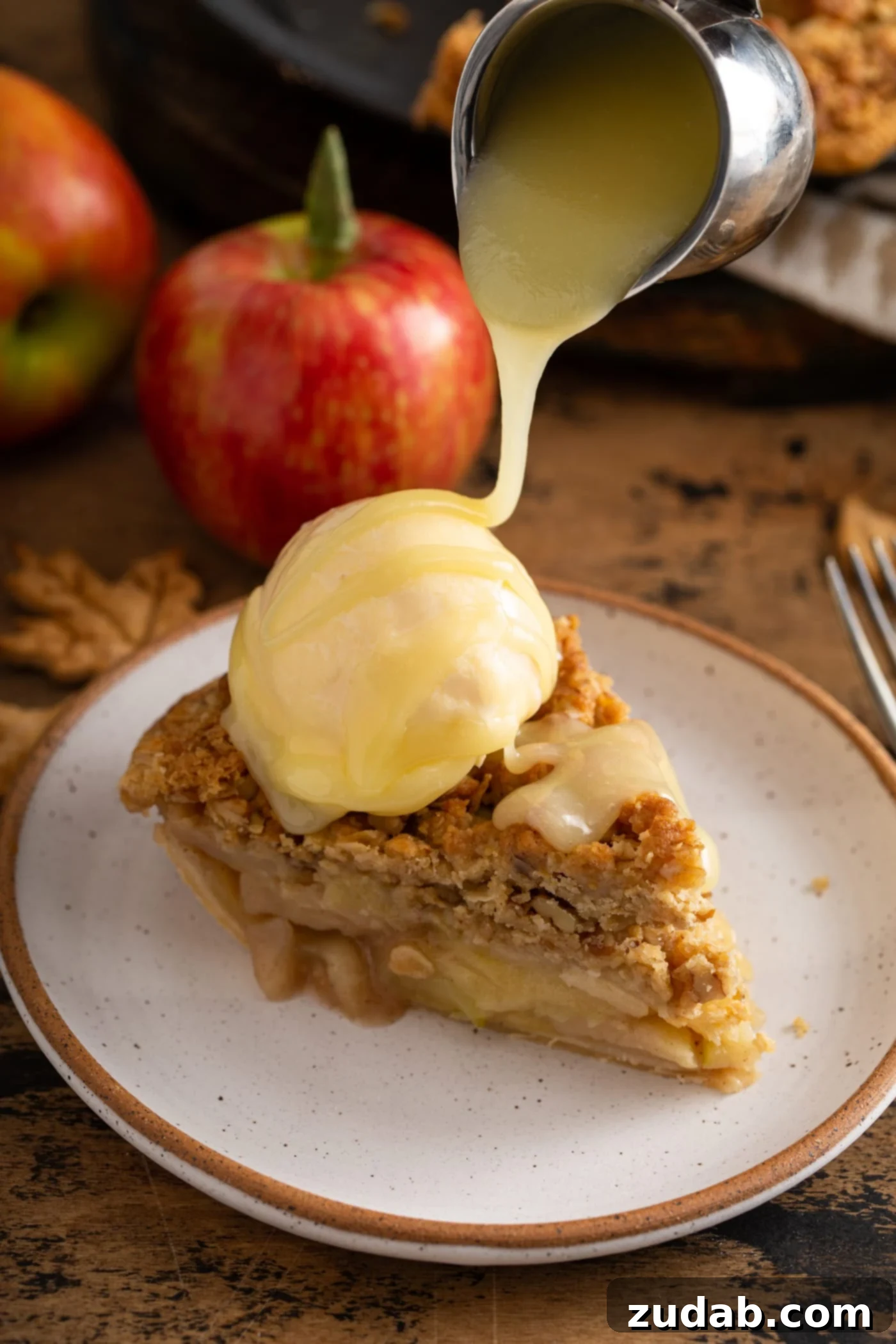 A generous slice of Dutch apple pie, topped with a scoop of vanilla ice cream, and decadently drizzled with rich white chocolate butter sauce. A vibrant red apple rests in the soft-focus background.