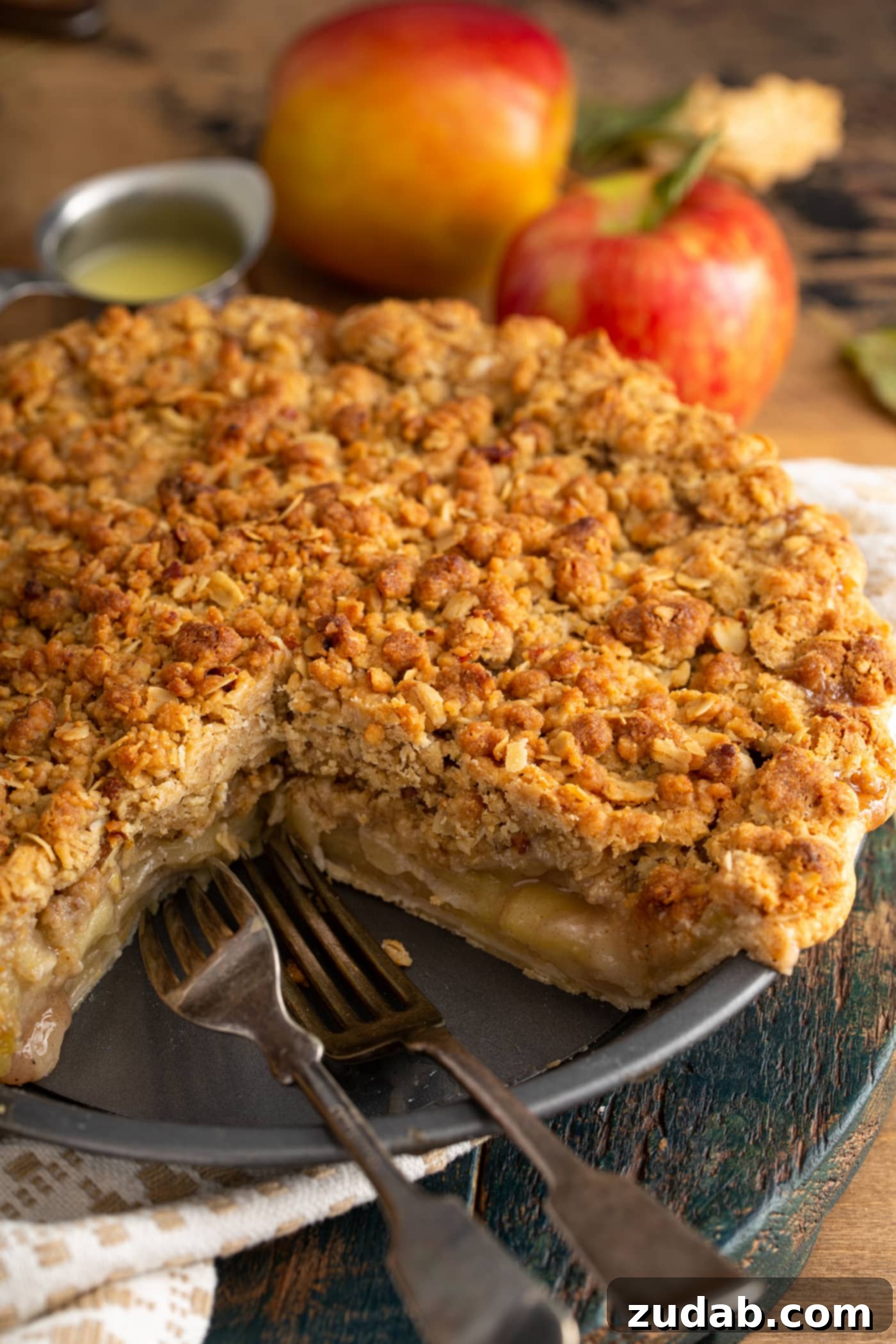 A freshly baked Dutch apple pie with one slice removed, showcasing the beautifully cooked apple filling and the golden-brown streusel topping. Two whole apples are in the background, hinting at the key ingredient.