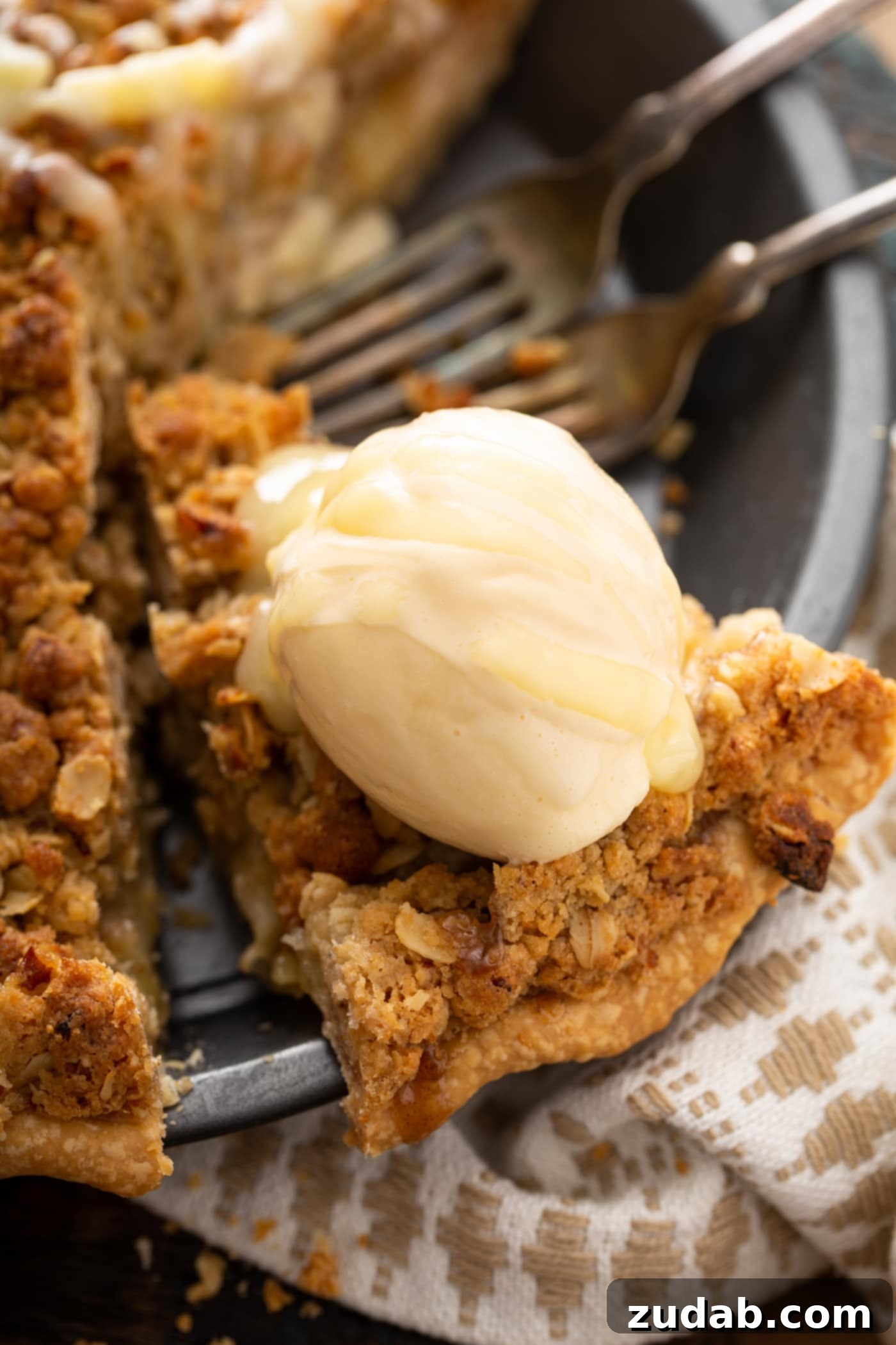 A hand lifting a slice of Dutch apple pie from the pie dish, revealing a scoop of melting vanilla ice cream and a luscious drizzle of white chocolate butter sauce cascading over it.