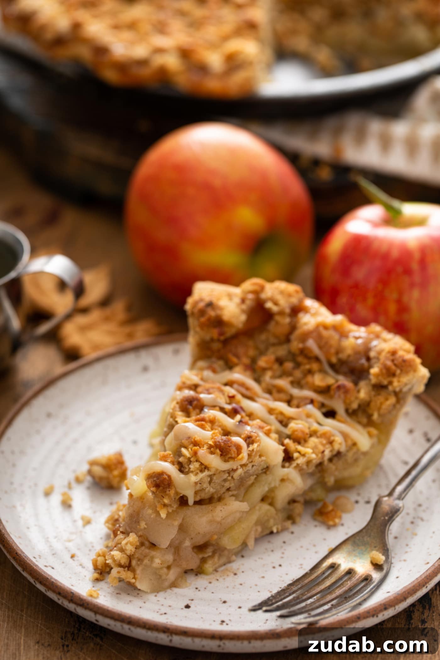 A slice of Dutch apple pie, beautifully plated and drizzled with a generous amount of white chocolate butter sauce. Two fresh apples are positioned in the background.