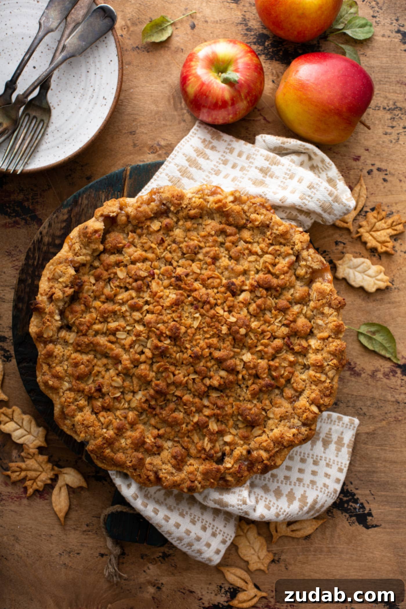 A freshly baked Dutch apple pie, boasting a beautifully golden-brown, crumbly streusel topping, sitting on a white tea towel. Fresh apples and a dessert plate are visible in the soft background.