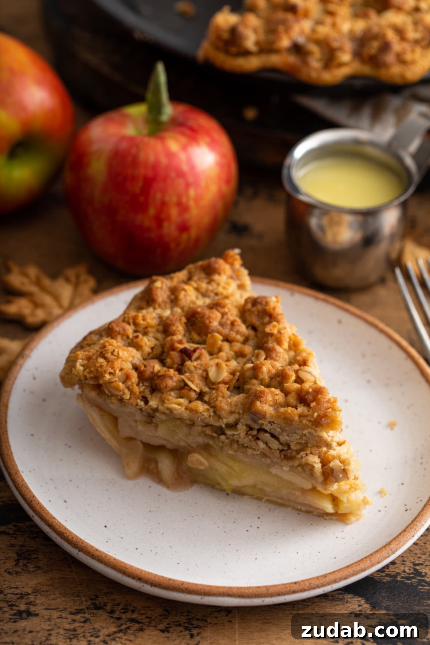A close-up of a slice of Dutch Apple Pie, revealing perfectly cooked apple slices nestled under a rich, golden-brown crumble topping. An apple and a container of the signature sauce are in the blurred background.