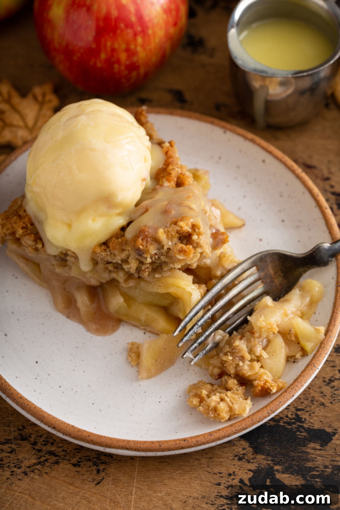 A delectable slice of Dutch apple pie on a white dessert plate, adorned with a scoop of vanilla ice cream and generously drizzled with warm white chocolate butter sauce. A fork is neatly placed beside the slice.