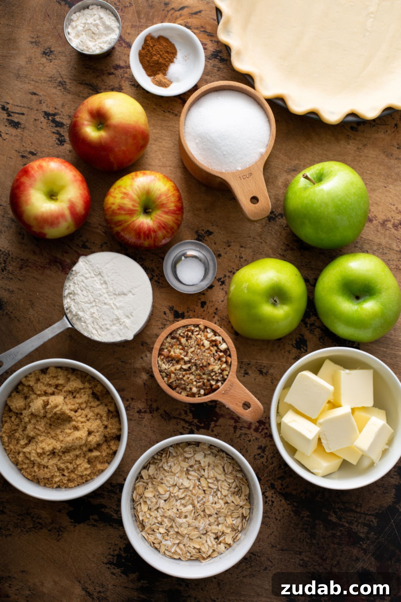 A collection of ingredients for Dutch Apple Pie, including green Granny Smith apples, red Honeycrisp apples, rolled oats, sticks of butter, granulated sugar, brown sugar, and a small dish of spices like cinnamon and nutmeg.