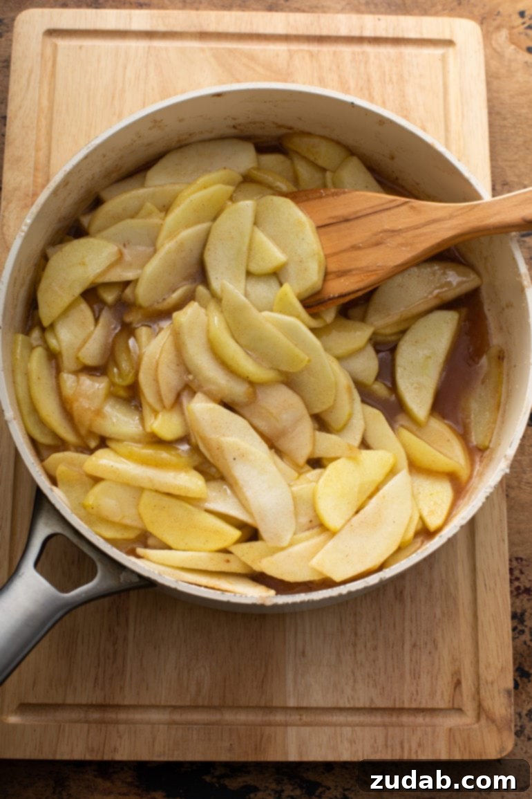 A stainless steel saucepan filled with sliced apples, being stirred with a wooden spoon into melted butter and sugar, a key step in preparing the pie filling.