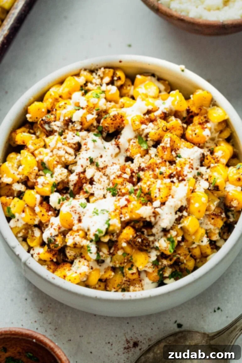 Oven Baked Sheet Pan Mexican Street Corn 6 A white serving bowl filled with perfectly roasted Mexican street corn, beautifully topped with crumbled cotija cheese, a dusting of chili powder, and fresh green cilantro sprigs.