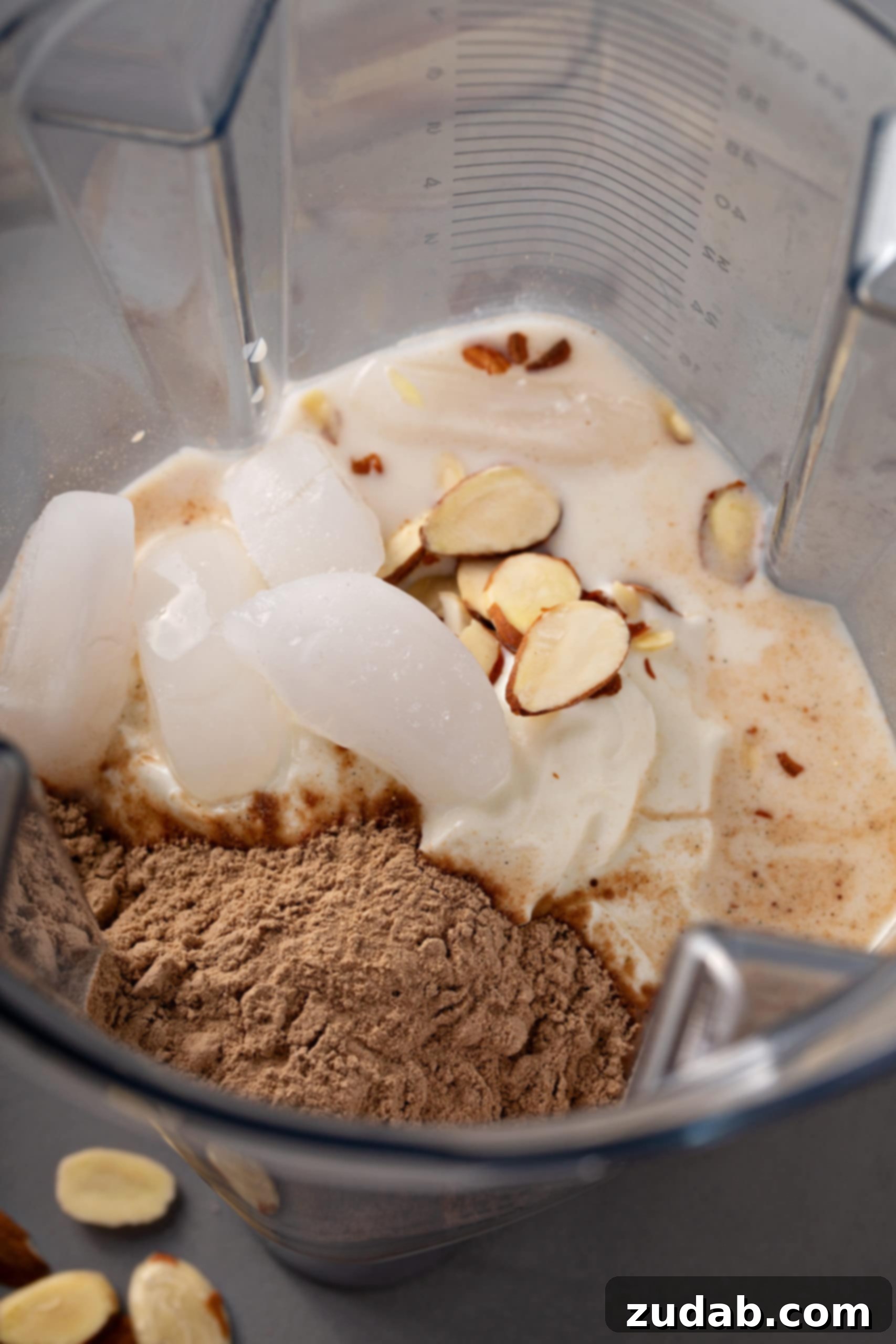 ingredients in a blender to make a chocolate coconut protein shake, ice cubes, almonds, chocolate protein powder, yogurt and milk