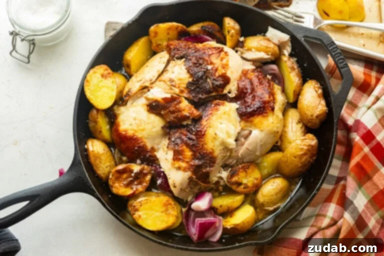 Garlic Butter Roasted Chicken and Crispy Potatoes 3 A whole roasted chicken with golden, crispy skin and a portion of the breast carved, revealing the succulent meat, surrounded by beautifully roasted potatoes in a large cast iron skillet.