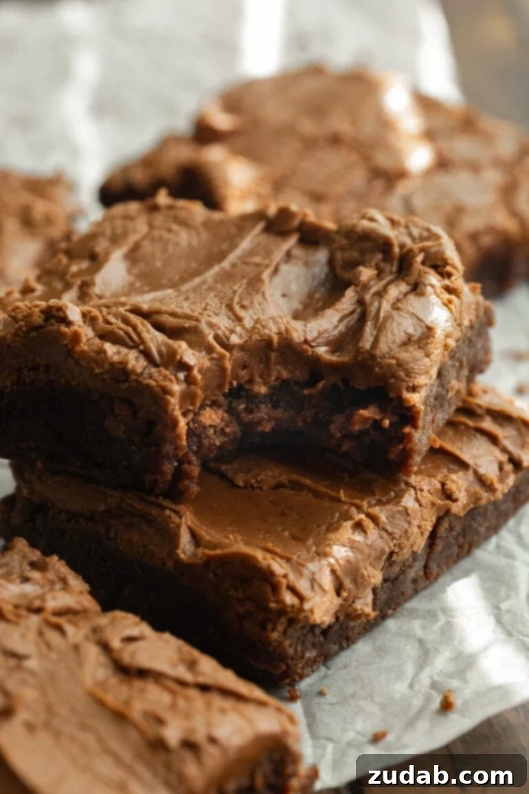 Classic Cafeteria Frosted Brownies 6 A mesmerizing close-up of deeply fudgy, frosted Lunch Lady Brownies, artfully stacked on parchment paper, revealing their gooey interiors and glossy chocolate topping.