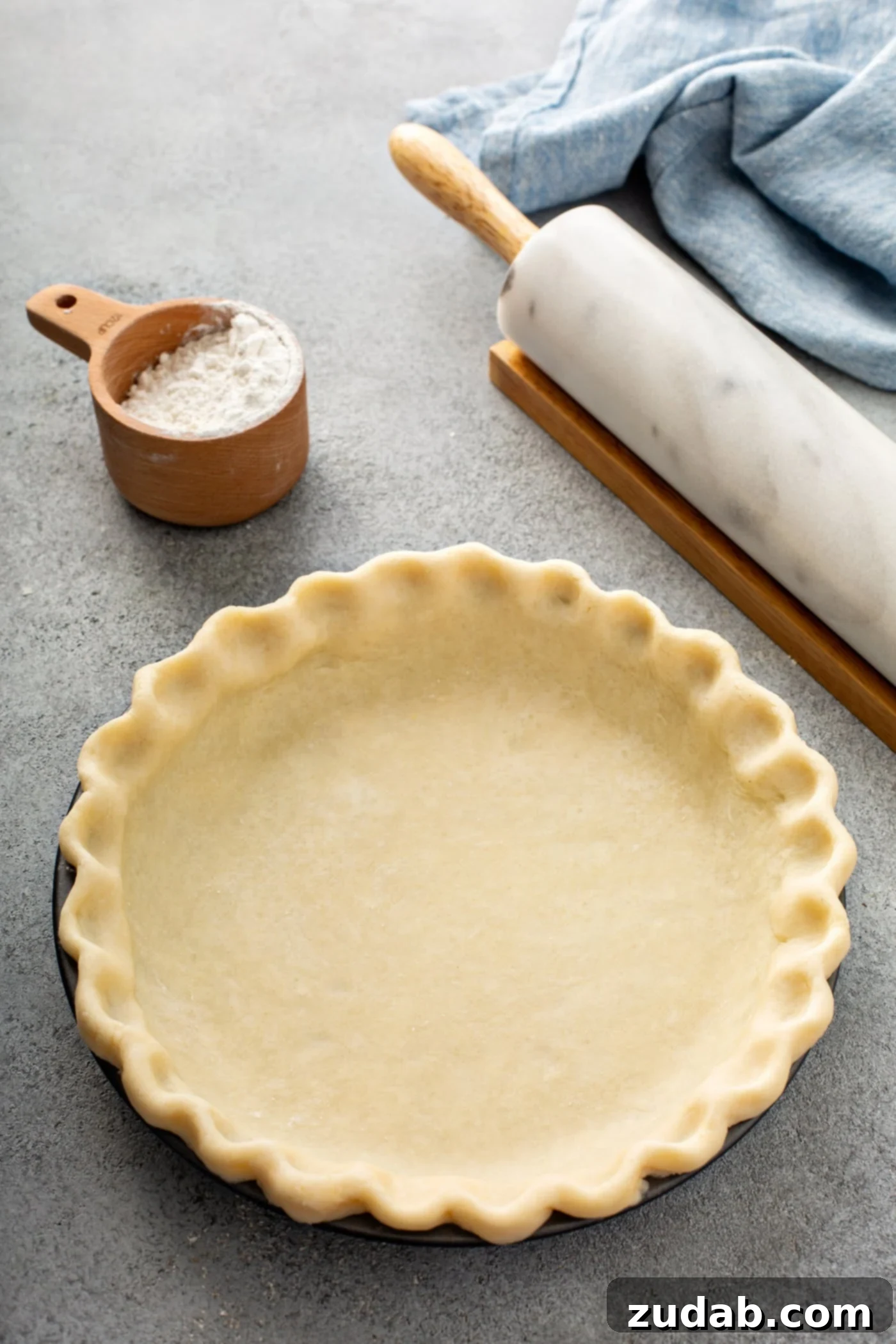 My Absolute FAVORITE Pie Crust Recipe A photo of an uncooked butter pie crust in a pie tin sitting on a table next to a rolling pin and a cup of flour.