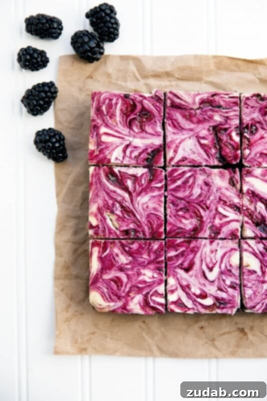 Artful Swirl: Blackberry Cheesecake Brownies That Look as Good as They Taste A photo taken over the top of blackberry swirled cheesecake brownies sitting on a piece of brown parchment paper with whole blackberries scattered around it, showcasing the rich colors and textures.