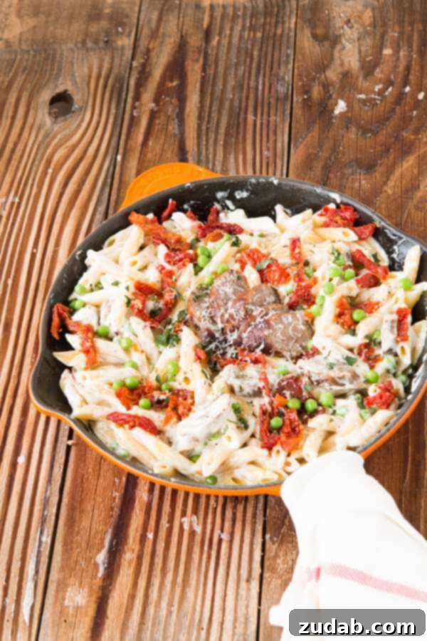 Hearty Sausage Alfredo Pasta with Sun-Dried Tomatoes Sausage Alfredo pasta in a large pan, tossed with sun-dried tomatoes and peas, capturing the rich texture of the creamy sauce