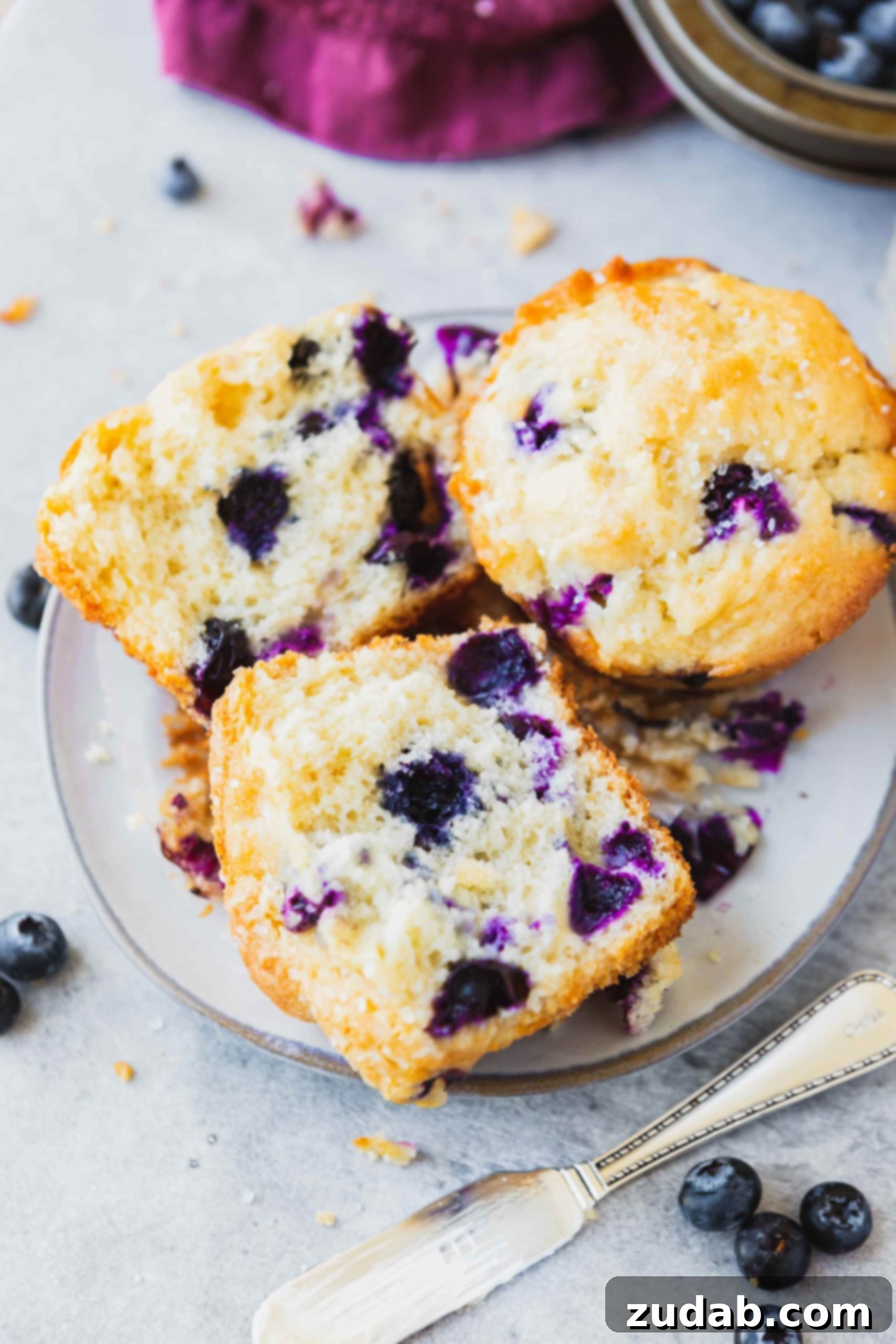 Two blueberry muffins on a small plate. One muffin is broken in half exposing the many blueberries inside.
