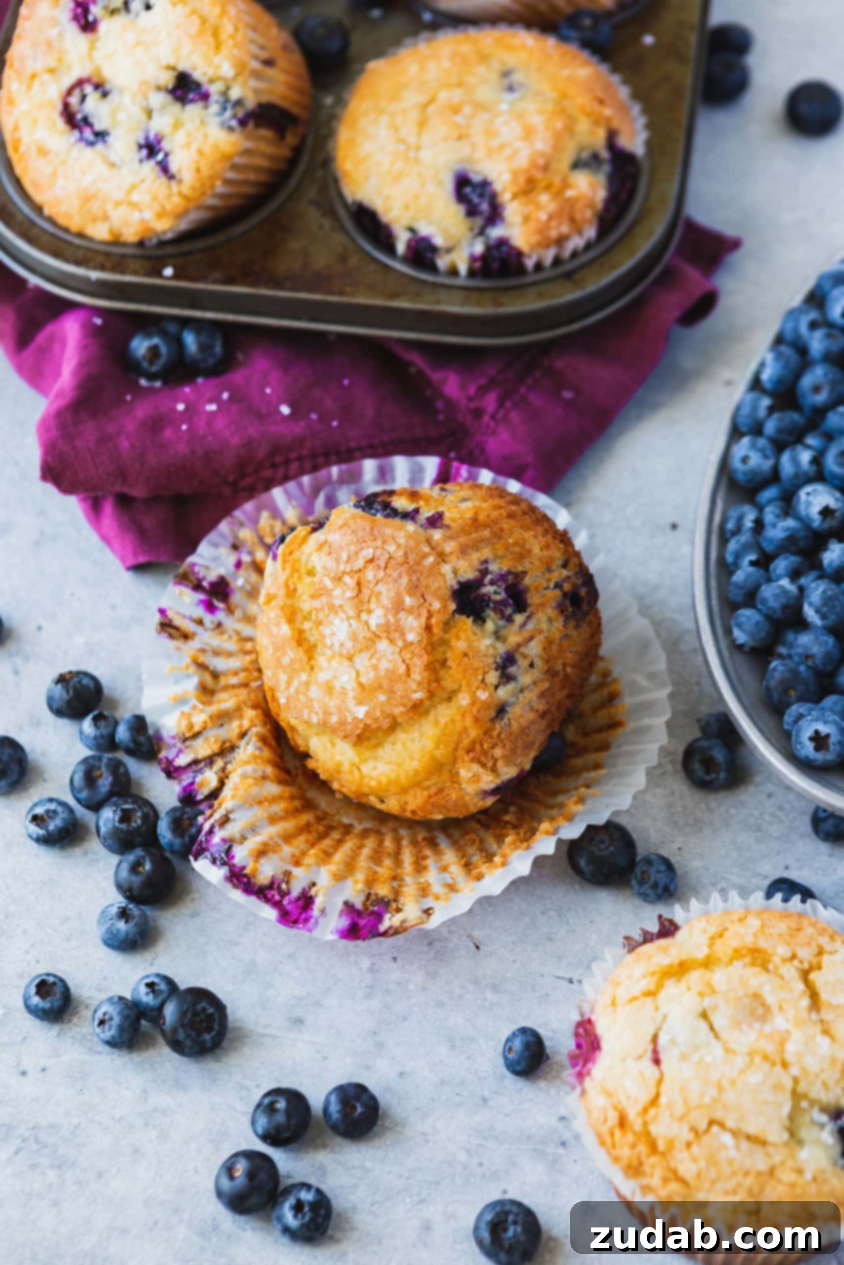 A blueberry muffin with the paper liner peeled down. There are blueberries showing in the muffin and blueberries on the table next to the muffin. A muffin tin with other muffins in the background.