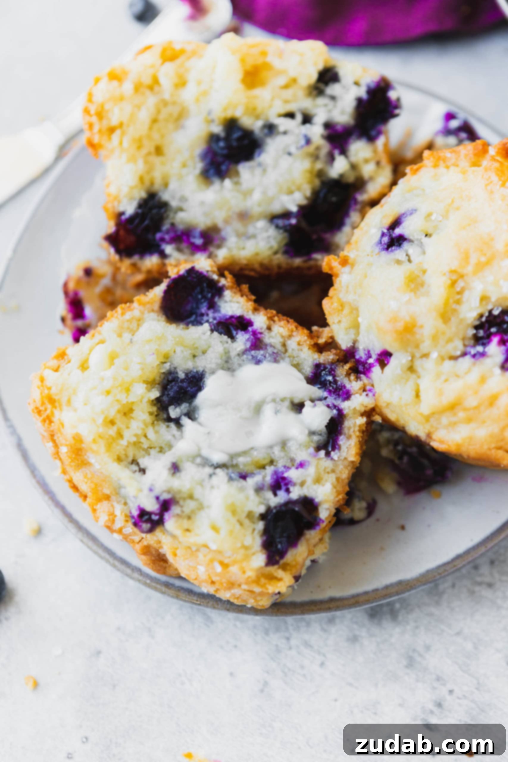 Two blueberry muffins on a small plate. One muffin is broken in half so you can see all of the blueberries packed inside.
