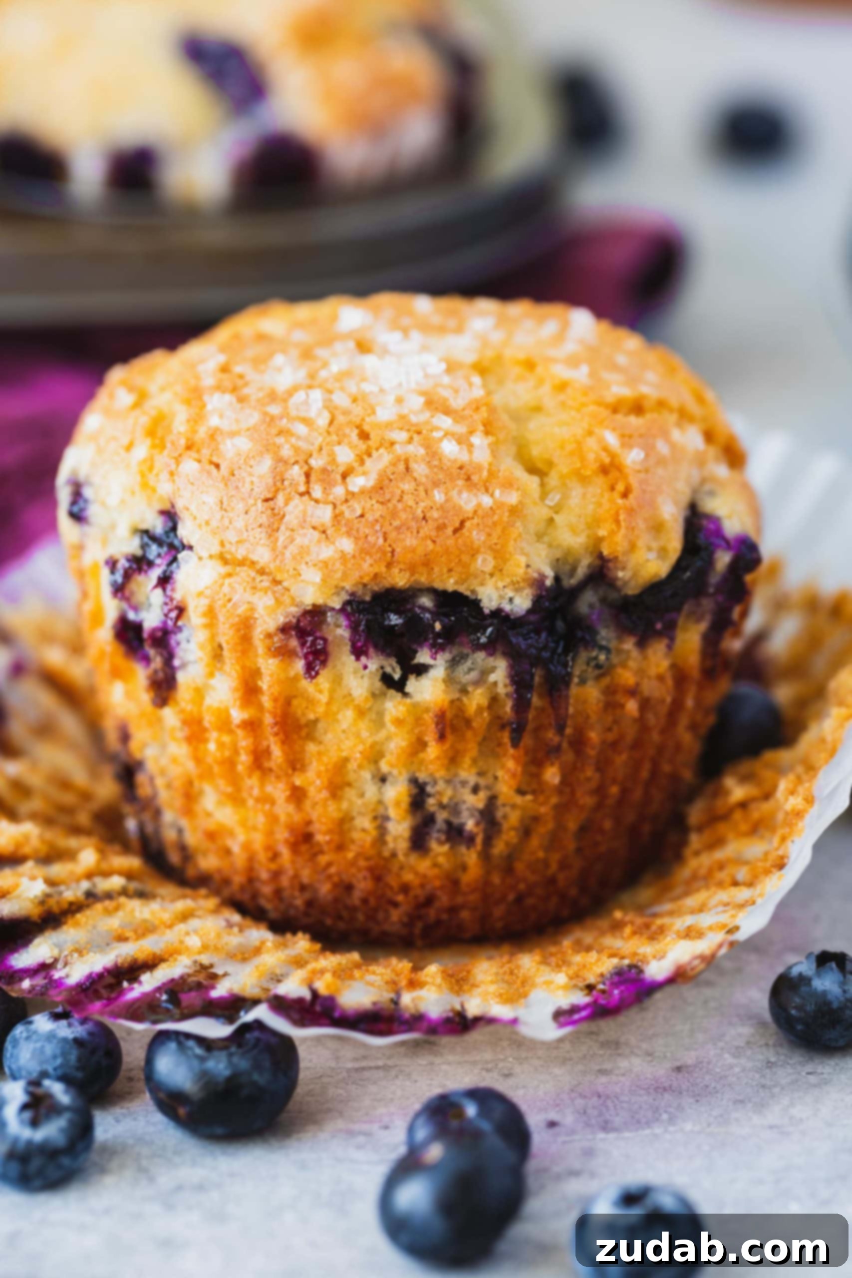 A blueberry muffin with the paper liner peeled back so you can see the golden brown muffin packed with blueberries.