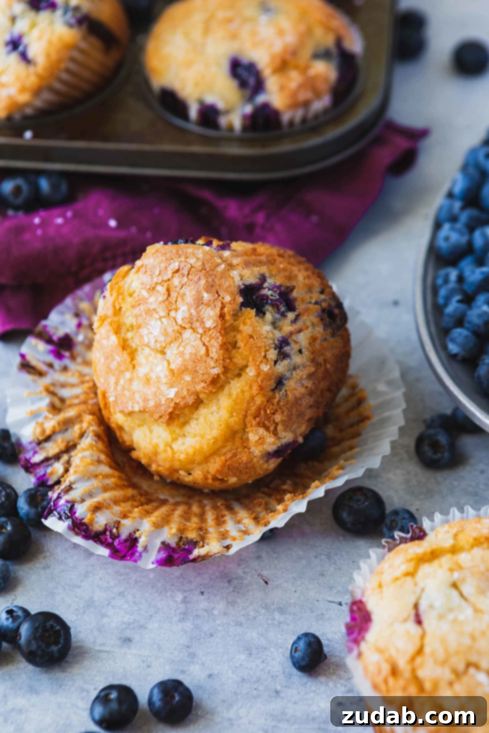 A blueberry muffin with the paper liner peeled down so you can see the baked sides of the muffin. There are blueberries in the muffin and on the table next to the muffin. there is another muffin and a bowl of blueberries in the background.