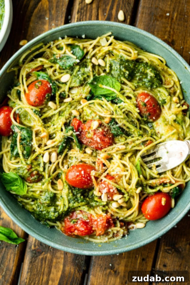 Quick Cheesy Pesto Pasta 2 A vibrant photo of cooked angel hair pasta tossed with fresh basil pesto, bright grape tomatoes, crunchy pine nuts, aromatic basil leaves, and grated Parmesan cheese, ready to be served.