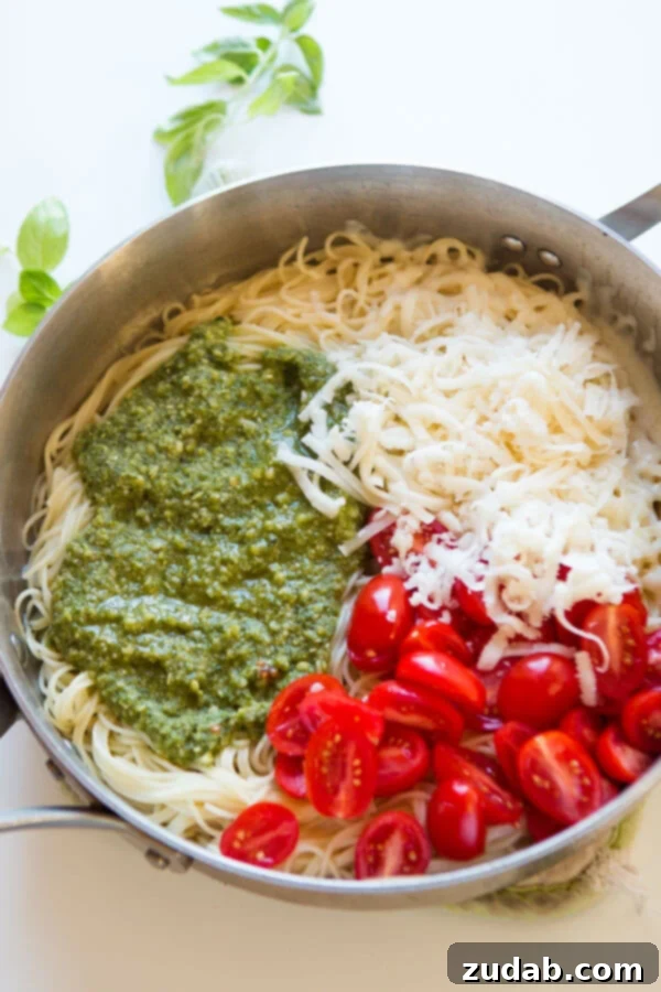Need dinner on the table in 20 minutes? We've got a 20 minute cheesy pesto pasta recipe that's quick and easy to make and it's a great special diets recipe. A close-up shot of key pesto pasta ingredients – fresh basil pesto, shredded mozzarella, cooked angel hair pasta, and vibrant grape tomatoes – beautifully arranged in a large skillet, ready for tossing.