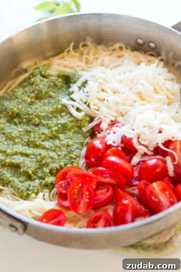 Need dinner on the table in 20 minutes? We've got a 20 minute cheesy pesto pasta recipe that's quick and easy to make and it's a great special diets recipe. A top-down view of the skillet, capturing the moment just before mixing, with luscious basil pesto, shredded mozzarella, al dente pasta, and juicy grape tomatoes waiting to be combined into a delectable dish.