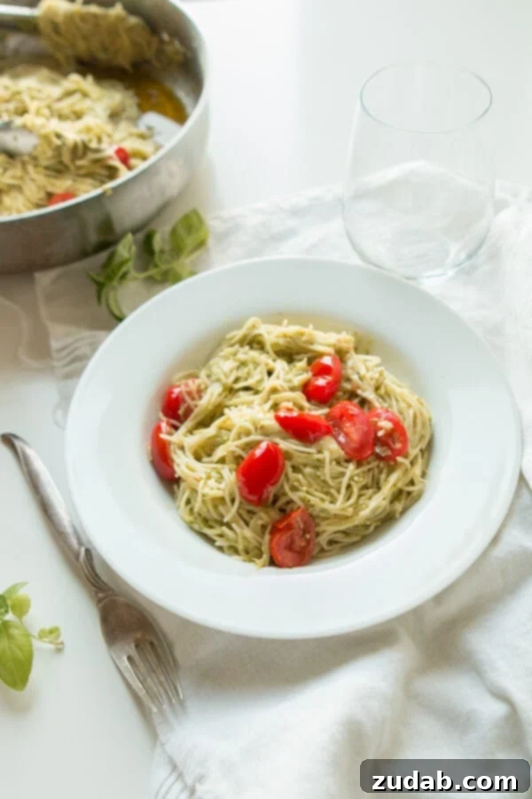 Need dinner on the table in 20 minutes? We've got a 20 minute cheesy pesto pasta recipe that's quick and easy to make and it's a great special diets recipe. A beautifully plated white bowl filled with vibrant basil pesto pasta, garnished with fresh basil leaves and grape tomatoes, sits beside a fork and a water glass on a clean white surface, highlighting its simple elegance.