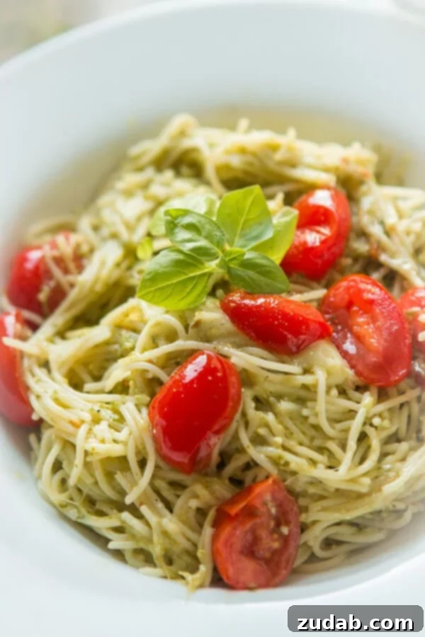 Need dinner on the table in 20 minutes? We've got a 20 minute cheesy pesto pasta recipe that's quick and easy to make and it's a great special diets recipe. A close-up of a white bowl showcasing a generous serving of pesto sauce pasta, beautifully garnished with fresh grape tomatoes and fragrant basil leaves, emphasizing its appetizing appeal.