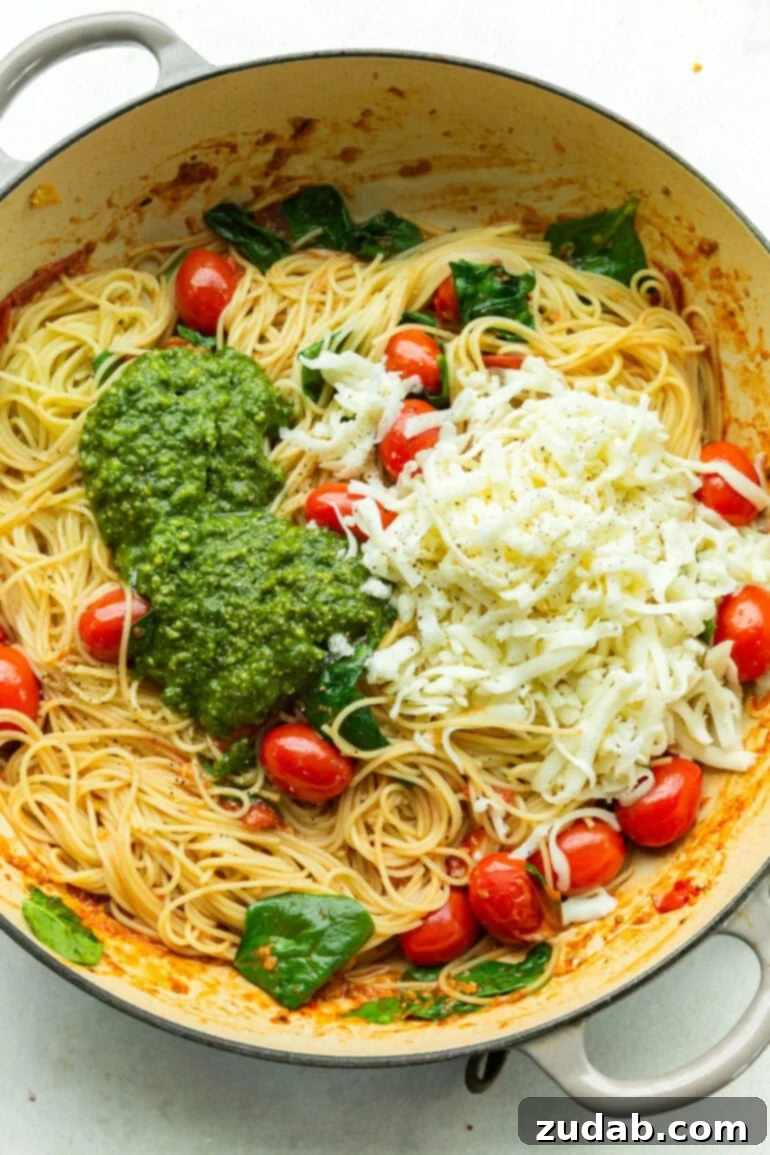 Quick Cheesy Pesto Pasta 3 A close-up photo showcasing cooked angel hair pasta in a large pot, generously topped with vibrant green pesto sauce, shredded mozzarella cheese, fresh basil leaves, and whole grape tomatoes, ready for a delicious toss.