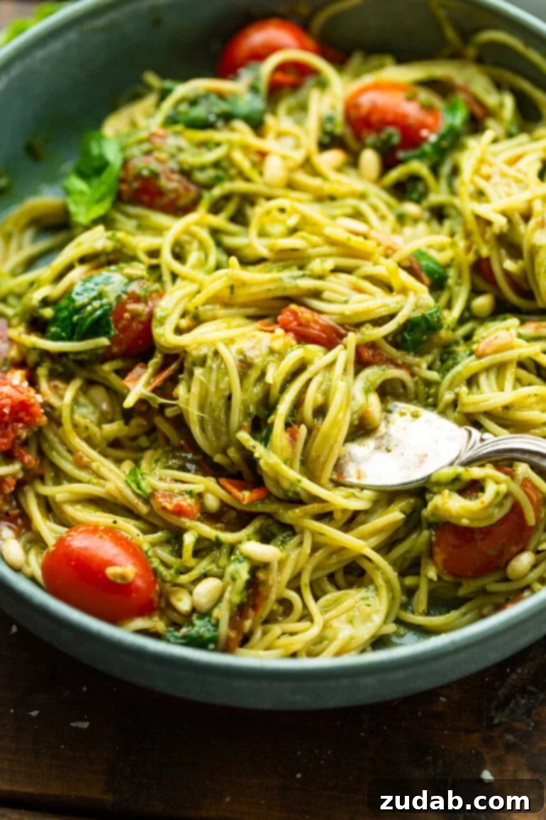 Quick Cheesy Pesto Pasta 7 A fork lifts a generous portion of pesto-covered angel hair pasta from a large, inviting bowl, showcasing the rich green sauce, melted cheese, and tender tomatoes, ready to be enjoyed.