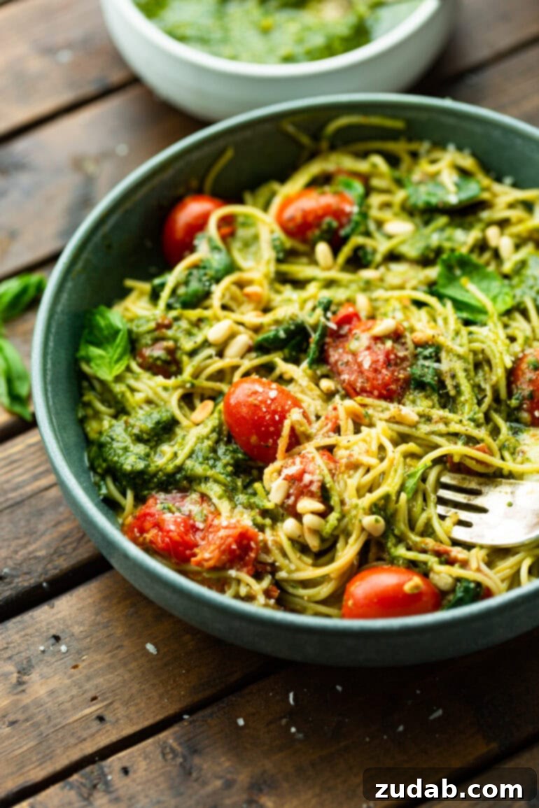 Quick Cheesy Pesto Pasta 9 A large, inviting bowl filled with cooked angel hair pasta, generously tossed with vibrant green pesto, juicy grape tomatoes, fresh basil leaves, melted mozzarella cheese, and crunchy pine nuts, ready for a delicious meal.