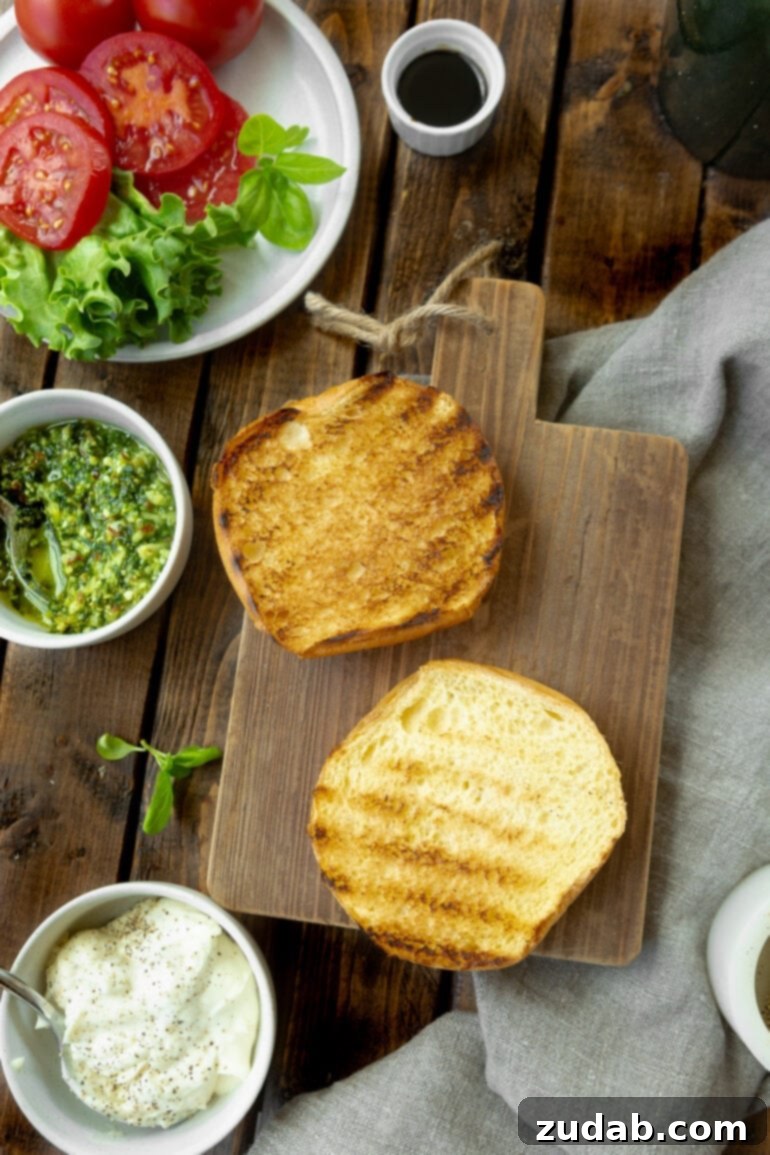 Aromatic Caprese Pesto Burgers 5 An open brioche bun, perfectly toasted with visible grill marks, rests face up on a wooden cutting board. Alongside, a small bowl of fresh pesto and a plate of vibrant lettuce and sliced tomatoes await, signifying the assembly of a gourmet burger.