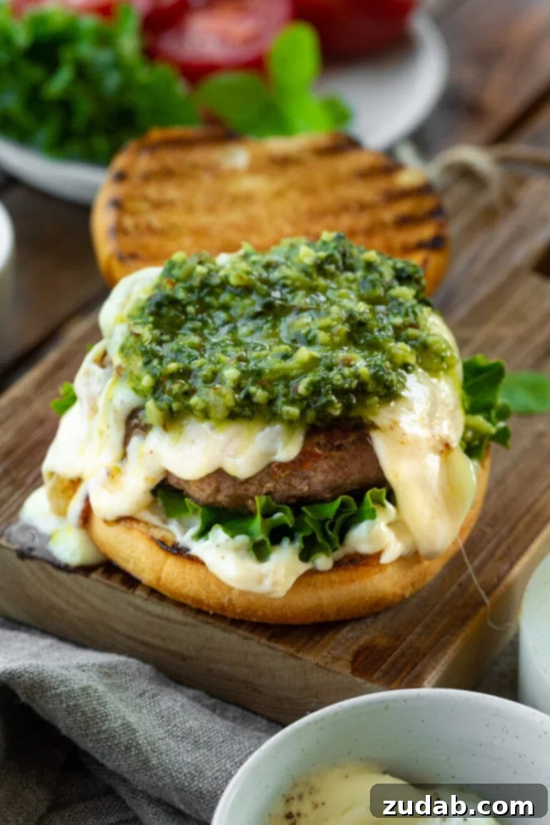 Aromatic Caprese Pesto Burgers 6 An exquisite burger with the top bun removed, revealing layers of creamy garlic aioli, fresh green leaf lettuce, perfectly melted Havarti cheese, and a vibrant green pesto topping. A testament to bold and delicious burger flavors.