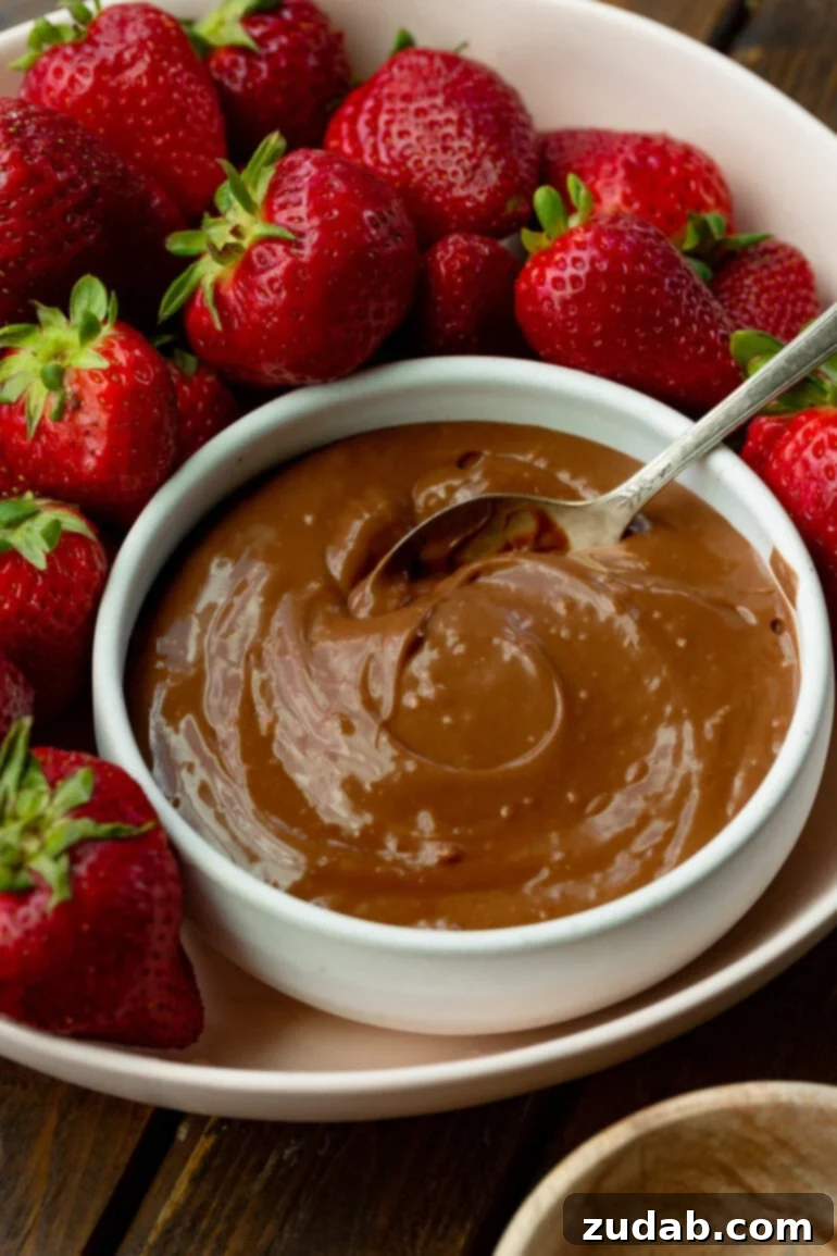 Guilt-Free Chocolate Strawberry Bliss 5 A small bowl full of a creamy chocolate dip, being stirred gently with a spoon, with fresh whole strawberries scattered around it on a white surface.