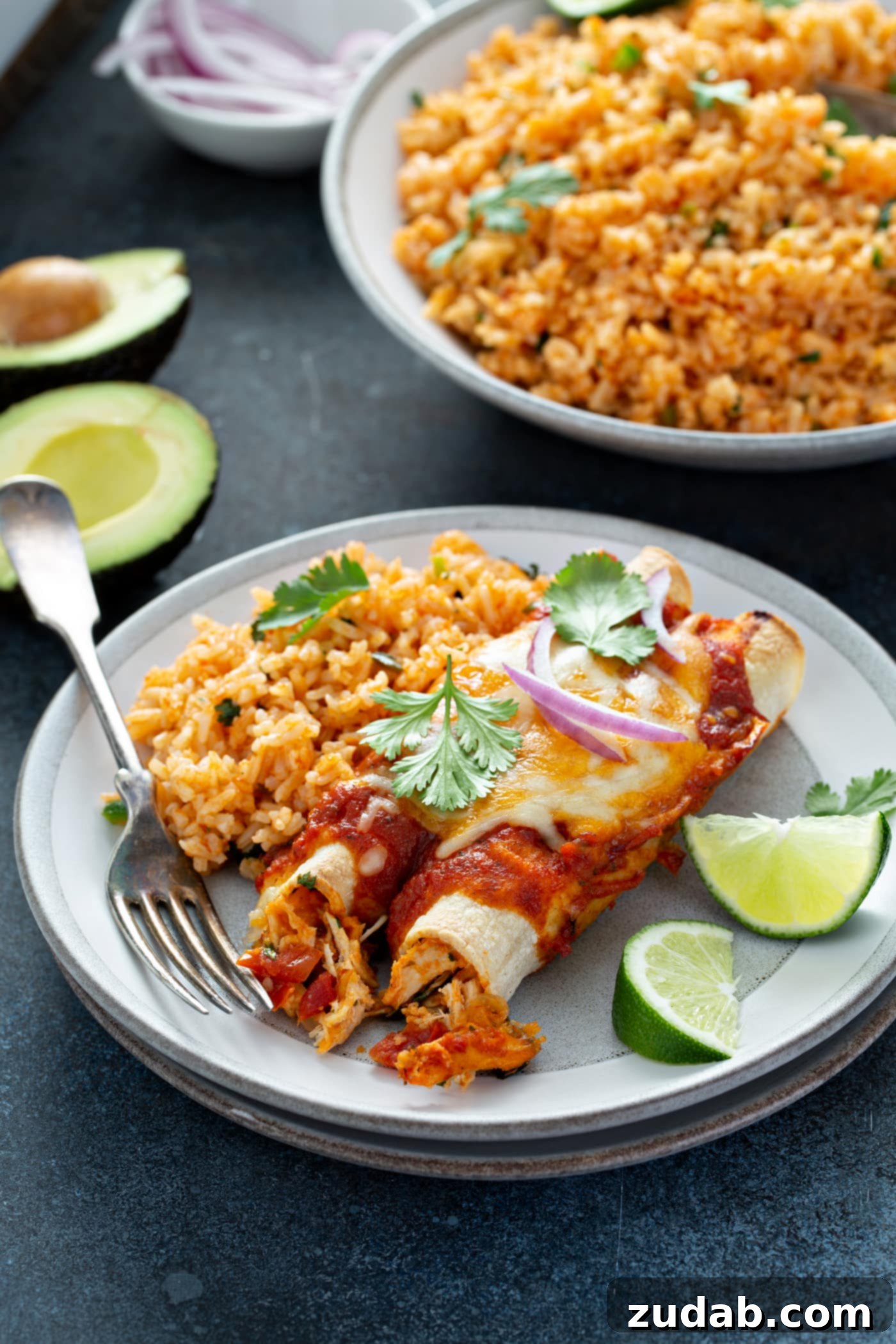 Red Chicken Enchiladas A serving plate with two enchiladas, Spanish rice, and two lime wedges. Cilantro leaves are sprinkled on top of the enchiladas. There is a bowl of Spanish rice and an avocado that is cut in half in the background.