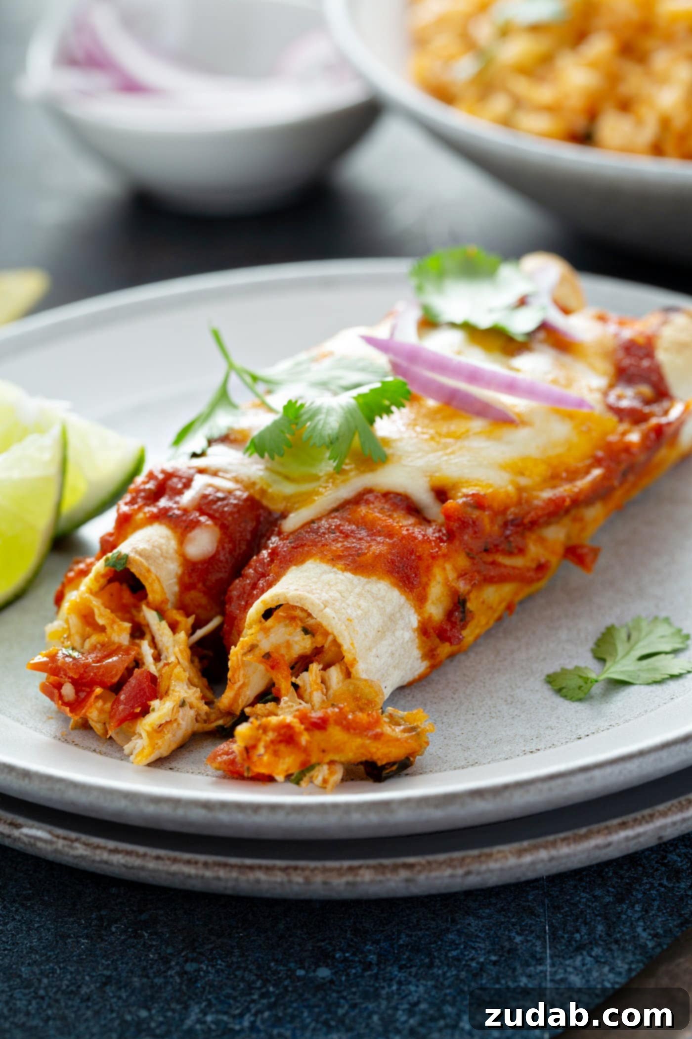 Red Chicken Enchiladas Two red chicken enchiladas on a dinner plate with red sauce, melted cheese and cilantro leaves on top. Two lime wedges are on the plate next to the enchiladas.